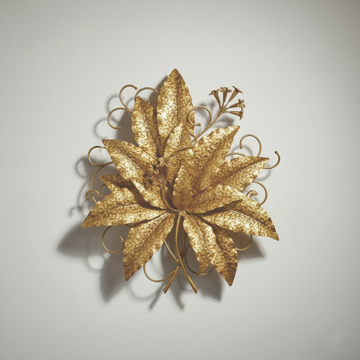 Close-up of a decorative, gold-finished metal wall sculpture featuring large, stylized tobacco leaves with a distinct hammered texture. The botanical piece is approximately 24"x24" and hangs against a white wall.