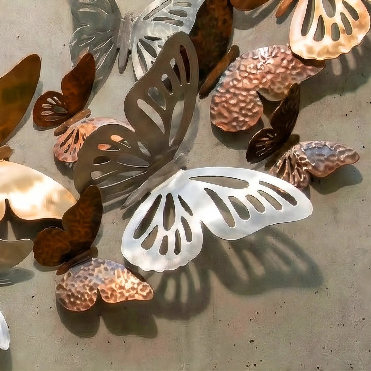Close-up detail of the Ascension metal wall sculpture, showing the layered arrangement and varied textures of hammered bronze, smooth gold, and brushed silver steel butterflies casting shadows on the wall.